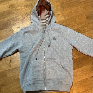 Grå hoodie från Burberry - Säljer en stilren grå hoodie från Burberry med dragkedja och klassiskt rutigt foder i huvan. Tröjan har en broderad logga på bröstet och praktiska fickor framtill. Perfekt för en avslappnad och trendig look.