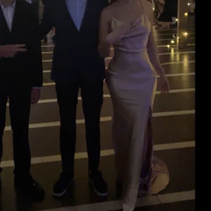 Champagnefärgad långklänning - Elegant champagnefärgad långklänning i satin med smala axelband och en stilren slits. Klänningen har en vacker omlott-design och en mjuk, glansig yta som ger en lyxig känsla.