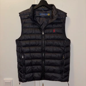 Svart dunväst från Polo Ralph Lauren - Säljer en stilren svart dunväst från Polo Ralph Lauren. Västen har en dragkedja framtill och är dekorerad med det ikoniska röda logotypbroderiet på bröstet. Nypris 3595,00kr. Har endast använt ett par gånger.