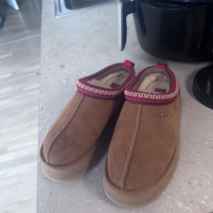 Bruna mockaskor från UGG - Säljer ett par bruna mockaskor från UGG med dekorativ kant i rött och vitt. Skorna har en robust sula och är fodrade för extra värme. Perfekta för kyligare dagar.