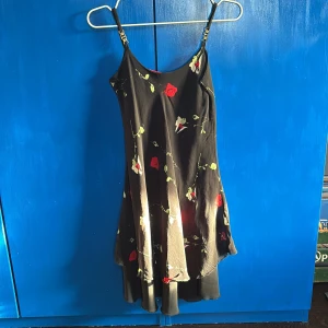 Svart blommig klänning med smala axelband - Säljer en så fin svart klänning med blommigt mönster i rött och grönt. Klänningen har smala axelband och en avslappnad passform. Det finns ingen lapp på den men den är köpt vintage men har aldrig använt den! ✨