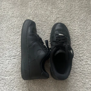 Svarta Nike Air Force 1 sneakers - Snygga svarta Nike Air Force sneakers i skinn med klassisk design. Skorna har snörning och en perforerad tå för extra ventilation. Perfekta för en stilren look. 