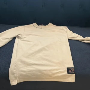 Vit långärmad tröja från Tommy Hilfiger - Säljer en stilren vit långärmad tröja från Tommy Hilfiger. Tröjan har en diskret logga vid halsen och en patch med flaggor och text på sidan. Perfekt för en avslappnad och snygg look.