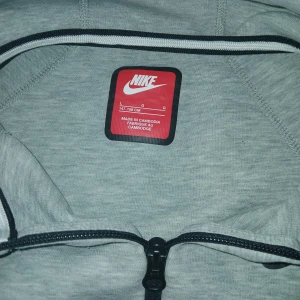 Grå huvtröja från Nike - Säljer en grå huvtröja från Nike med dragkedja och två fickor framtill. Nike-loggan tryckt på bröstet. Skick-8/10 inte använd så många gånger. Nypris- 1200 i butik där den var köpt. Storlek finns på bilden men passar otroligt bra på mig som är 160 cm.