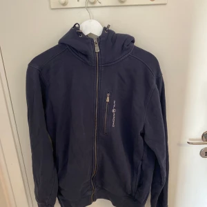 Mörkblå hoodie från Sail Raicing - Säljer en mörkblå hoodie från Sail Raicing med dragkedja och huva. Jackan har en bröstficka med dragkedja och långa ärmar. Perfekt för kyliga dagar.