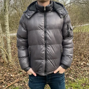 Grå pufferjacka från Brave Soul - Säljer en grå pufferjacka från Brave Soul. Jackan har en hög krage och en huva för extra värme. Den är utrustad med dragkedja och har praktiska fickor på sidorna och en på ärmen. Perfekt för kyliga dagar. Köpt på Zalando nypris 979kr