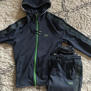 Svart hoodie från Hugo Boss - Snygg svart hoodie från Hugo Boss med gröna detaljer och dragkedja. Den har långa ärmar och en huva med snörning. Logotypen finns på bröstet och längs ärmarna. Perfekt för en stilren och bekväm look. 