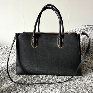 Svart handväska - Svart handväska med praktiska handtag och avtagbar axelrem. Väskan har en stilren design med guldfärgade dragkedjor och rymligt inre för alla dina nödvändigheter. Endast anvånd nån gång och har inga tecken på användning.