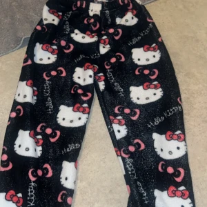 Svarta Hello Kitty mjukisbyxor - Mysiga svarta mjukisbyxor med Hello Kitty-motiv i vitt och rosa. Perfekta för en avslappnad dag hemma. Byxorna har en bekväm passform och är gjorda i ett mjukt material.