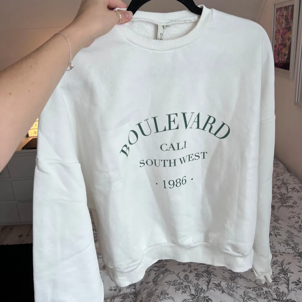 Säljer en vit sweatshirt med texten 'Boulevard Cali South West 1986' tryckt på framsidan. Tröjan är oversized i storleken, har långa ärmar och ribbade muddar vid ärmslut och nederkant. Perfekt för en avslappnad stil. Önskar du veta exakta mått? Tveka inte att höra av dig!. Neuletakit & Villapaidat.