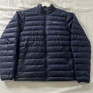 Mörkblå dunjacka från Patagonia - Säljer en snygg mörkblå dunjacka från Patagonia. Jackan har en dragkedja framtill och två praktiska fickor med dragkedjor. Perfekt för kyliga dagar med sin quiltade design och långa ärmar. En stilren och funktionell jacka för alla tillfällen. Den är i bra skick och ger ett riktigt bra helhetsintryck. Är storlek S men passar även M