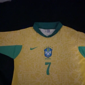 Brasilien/fotbollströja - Säljer en gul Brasilien fotbollströja från Nike med gröna detaljer och nummer 7. Tröjan har korta ärmar och ett tryck för Copa America USA 2024 på ärmen. Perfekt för fotbollsfans! Tröjan är liten i storleken, kom privat för mer information, helt ny