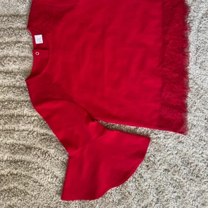 Röd blus från Lindex Kids (128) - Säljer en röd blus från Lindex Kids med vackra spetsdetaljer och volangärmar. Perfekt för en färgglad look. Blusen har en rund hals och knappar i ryggen för enkel påklädning.