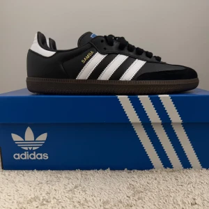 Adidas Samba OG - Klassiska Adidas Samba OG sneakers i svart och vitt med de ikoniska tre ränderna på sidan. Skorna har en brun sula och är perfekta för en stilren look. De är märkta med 'SAMBA' i guld på sidan.