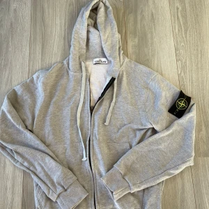 Grå hoodie från Stone Island - Säljer en stilren grå hoodie från Stone Island med dragkedja och justerbar huva. Tröjan har en ikonisk logga på ärmen och ribbade muddar vid ärmslut och nederkant. Perfekt för en avslappnad look.