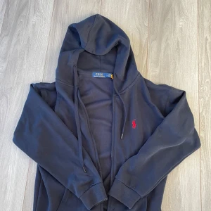Mörkblå hoodie från Ralph Lauren - Säljer en snygg mörkblå hoodie från Ralph Lauren med det klassiska röda logotypbroderiet på bröstet. Tröjan har en dragkedja framtill och justerbara snören i huvan. Perfekt för en avslappnad stil. !OBS! (Storleken är en large men passar som en small)