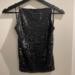 Svart paljettlinne - Snyggt svart linne täckt med glittrande paljetter. Linnet har smala axelband och en figurnära passform som ger en festlig look. Perfekt för att sticka ut på festen!