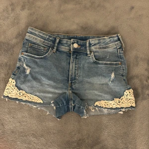 Jeansshorts med spetsdetaljer från H&M - Snygga jeansshorts från H&M med slitna detaljer och spetsapplikationer vid benen. De har en klassisk femficksdesign och knappgylf.