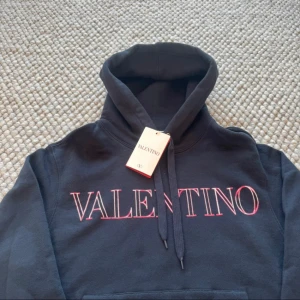 Svart hoodie från Valentino - Snygg svart hoodie från Valentino med rosa logotyp på bröstet. Den har en klassisk design med justerbar huva och en stor magficka framtill. Perfekt för en stilren och bekväm look.