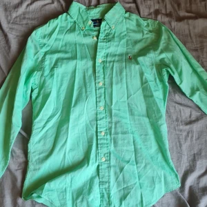Grön skjorta från Ralph Lauren - Snygg grön skjorta från Ralph Lauren i slim fit. Skjortan har långa ärmar och knappar framtill. Den är broderad med den klassiska Ralph Lauren-loggan på bröstet. Perfekt för en stilren look!  Priset är diskuterbart