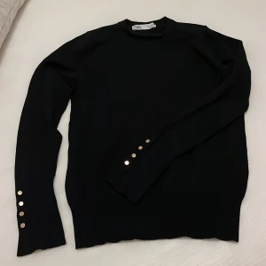 Svart tröja från Zara med knappar - Säljer en stilren svart tröja från Zara med långa ärmar och dekorativa knappar längs ärmsluten. Perfekt för en elegant look. Tröjan är i en klassisk design som passar till många tillfällen.