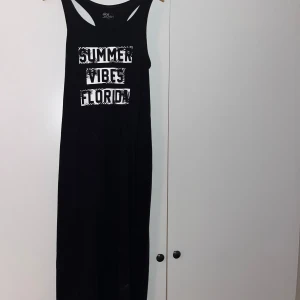 Svart långklänning från Ellos - Snygg svart långklänning från Ellos med texten 'Summer Vibes Florida' i vitt på framsidan. Perfekt för en avslappnad sommarstil. Klänningen är ärmlös och har en bekväm passform. Är stretchig och passar många storlekar🌸 skriv för fler bilder 