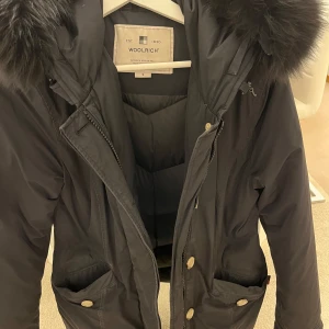 Svart dunjacka från Woolrich - Säljer en svart dunjacka från Woolrich med pälskantad huva och dragkedja. Jackan har praktiska fickor och en stilren design, perfekt för kyliga dagar. Den är både varm och bekväm, vilket gör den till ett måste i vintergarderoben. 🖤den har äkta päls och köptes från Johnells , den täcker baken o väldig varm och är i bra skick , den köptes för 10 000kr, säljes för endast 2 000kr. Pris kan diskuteras vid snabb affär 