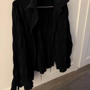 Svart blus med volangdetaljer - Snygg svart blus med volangdetaljer på ärmarna. Blusen har en krage och knappar framtill, vilket ger en stilren look. Perfekt för att lägga till en touch av elegans i din outfit.