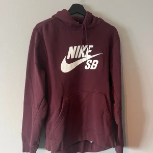 Vinröd hoodie från Nike SB - Säljer en snygg vinröd hoodie från Nike SB med stor logga på framsidan. Tröjan har en klassisk känguruficka och justerbar huva med dragsko. Perfekt för en avslappnad stil.