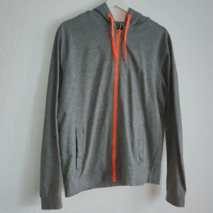 Grå hoodie med Dragon Ball-tryck från Roly - Säljer en grå hoodie från Roly med ett färgglatt Dragon Ball-tryck på ryggen. Hoodien har en dragkedja och orangea snören vid luvan. Perfekt för fans av serien! 🐉