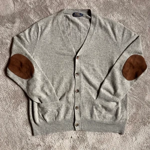 Grå Ralph lauren cardigan - Säljer en stilren grå kofta från Polo Ralph Lauren med bruna armbågslappar. 100% merinoull vilket gör den väldigt bekväm. I storlek XL men passar även M/L
