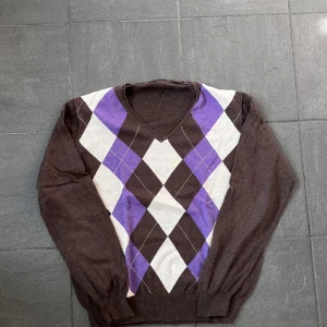 Brun tröja med argylemönster från Massimo Dutti - Säljer en stilren brun tröja från Massimo Dutti med ett klassiskt argylemönster i lila och vitt. Tröjan är tillverkad i en mjuk blandning av bomull och kashmir, perfekt för kyligare dagar. Den har långa ärmar och en v-ringad hals. Passar både till vardags och mer uppklädda tillfällen.