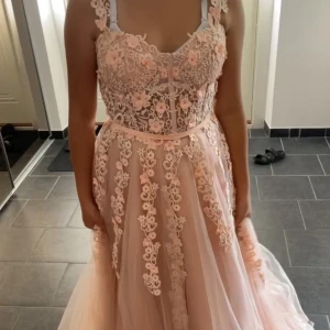 Rosa spetsklänning med blommor - Underbar rosa långklänning med spetsdetaljer och blommönster. Klänningen har en vacker öppen rygg och axelbandslös design med en elegant rosett i midjan. Perfekt för speciella tillfällen.