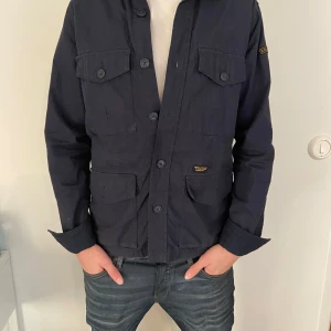 Overshirt - Snygg mörkblå jacka från Superdry med knappar framtill och flera praktiska fickor. Jackan har långa ärmar och en klassisk krage. Perfekt för en stilren look.Den är i mycket bra skick men inga defekter.