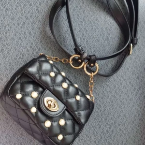 Svart quiltad axelväska med pärlor - Snygg svart quiltad axelväska med pärldetaljer och guldfärgad kedja. Väskan har en elegant vridlåsdetalj framtill och justerbar axelrem. Perfekt för att ge en touch av glamour till din outfit.