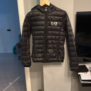 Svart pufferjacka från Emporio Armani - Snygg svart pufferjacka från Emporio Armani med EA7-logga på bröstet. Jackan har en dragkedja framtill och är quiltad för extra värme. Perfekt för kyligare dagar med sin långa ärmar och huva. Men syd vid mitten 
