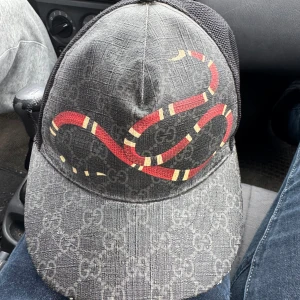 Svart keps med orm från Gucci - Snygg svart keps från Gucci med ett coolt ormmotiv i rött och gult. Kepsen har ett diskret mönster med märkets logotyp och ett nätmaterial på baksidan för extra ventilation. Perfekt för att ge din outfit en lyxig touch.  ÄKTA KEPS 
