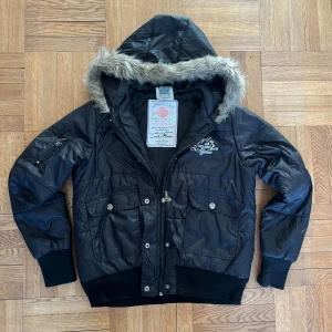 Svart dunjacka med pälsdetaljer - Snygg svart dunjacka från Salty Dog med pälsfodrad huva och coolt tryck på ryggen. Jackan har dragkedja och knappar framtill samt flera praktiska fickor. Perfekt för kyliga dagar när du vill hålla stilen. passar både s och m