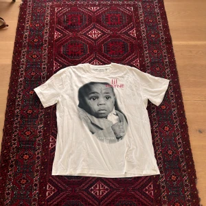 Lil Wayne Tshirt (Tryck Tshirt) - Säljer en vit t-shirt från Lil Wayne med stort svartvitt tryck av ett barn på framsidan och röd text. T-shirten har rund hals och är kortärmad med loose fit. Perfekt för dig som gillar streetwear och musikmerch.
