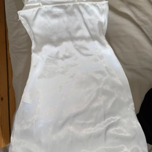 Vit satinklänning med smala axelband - Säljer en elegant vit satinklänning med smala axelband och figurnära passform. Klänningen har en enkel och stilren design som ger en lyxig känsla. Perfekt för speciella tillfällen eller när du vill känna dig extra fin.
