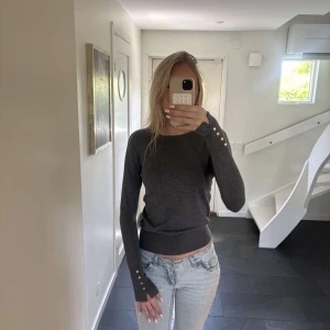 Mörkgrå tröja med gulddetaljer - Säljer en mörkgrå långärmad tröja med rund halsringning och dekorativa guldfärgade knappar vid ärmsluten. Tröjan har en smal passform och är perfekt för en stilren vardagslook.