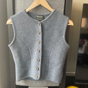 Grå stickad väst från Astrifa - Säljer en grå stickad väst från Astrifa med dekorativa knappar framtill. Västen har en rund halsringning och fina detaljer längs kanterna. Perfekt att styla över en skjorta eller tröja för en klassisk look.