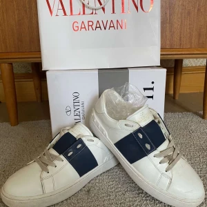 Valentino Garavani Open sneakers med blå detalj - Säljer ett par vita Valentino Garavani Open sneakers med bred mörkblå rem över mitten och silvriga nitar på sidorna. Skorna har rund tå, platt sula och vita skosnören. Klassisk och stilren design med exklusiv känsla.