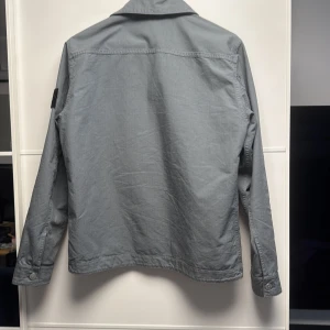 Grå overshirt från Stone Island - Säljer en stilren grå overshirt från Stone Island med klassisk krage och två stora bröstfickor. Jackan har lång ärm och ikonisk logotyp-patch på vänster arm. Perfekt för lager-på-lager och enkel att matcha med olika outfits.