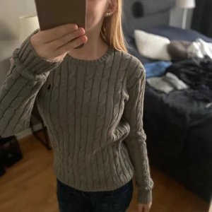 Grå kabelstickad tröja från Ralph Lauren - Säljer en klassisk grå kabelstickad tröja från Ralph Lauren med rund halsringning och diskret broderad logga på bröstet. Tröjan har lång ärm och är perfekt för lager-på-lager-stil. Supermjuk och stilren!