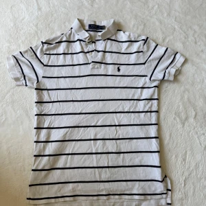 Vit randig pikétröja från Polo Ralph Lauren - Vit pikétröja från Polo Ralph Lauren med svarta horisontella ränder och klassisk krage. Tröjan har korta ärmar och liten broderad logga på bröstet. Perfekt för en stilren och avslappnad look.