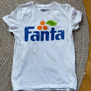 Vit Fanta t-shirt med tryck - Säljer en vit t-shirt med stort Fanta-tryck i blått, orange och grönt framtill. T-shirten har rund hals och korta ärmar. Perfekt för dig som gillar retrostil och färgglada loggor! Aldrig använd. Jag är 160cm
