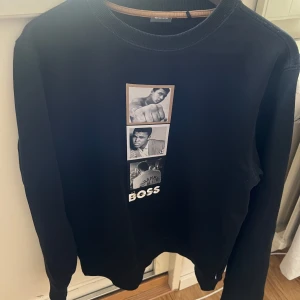 BOSS x Muhammad Ali - Svart sweatshirt från BOSS i samarbete med Muhammad Ali. Tröjan har tryck med bilder och texten 'BOSS' framtill samt tryck i nacken. Rund halsringning och långa ärmar. Perfekt för dig som gillar sportiga och stilrena plagg.