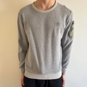 Grå sweatshirt från G-Star RAW - Säljer en grå sweatshirt från G-Star RAW med rund hals och ribbade muddar. Tröjan har en unik fickdetalj på ena ärmen i ljusare tyg. Perfekt för en avslappnad och stilren look. Skriv kring mer information😀