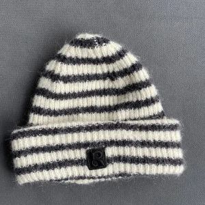 Randig stickad mössa från Rodebjer - Mysig ribbstickad mössa från Rodebjer i svart och vit med klassiskt randigt mönster. Tillverkad i en mjuk ull- och alpackablandning som håller dig varm under kalla dagar. Perfekt accessoar för att lyfta din vinterstil.